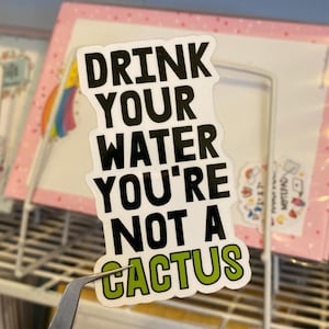 Könnte beinhalten: Ein weißer Aufkleber mit schwarzem Text, der "Drink your water you're not a cactus" lautet. Das Wort "cactus" ist in grüner Schrift. Der Aufkleber wird von einer Pinzette gehalten.