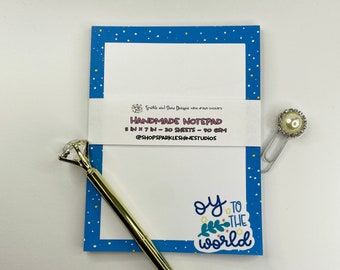 May include: A blue notepad with white polka dots and a sticker that says "oy to the world". The notepad is 5 inches by 7 inches and has 30 sheets. It is handmade and comes with a gold pen and a pearl-embellished paper clip.