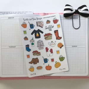 May include: A planner with a pink cover, open to a grid-lined page. A sheet of fall-themed stickers with illustrations of coffee cups, pumpkins, and boots is visible. A black and white striped bow clip and a pen are also present.