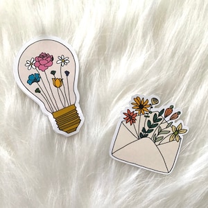 Può includere: Due adesivi con motivi floreali. Il primo adesivo è una lampadina con fiori colorati all'interno. Il secondo adesivo è una busta con un mazzo di fiori all'interno.