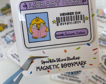 Puede incluir: Una tarjeta de biblioteca blanca y morada con un pollito amarillo leyendo un libro. La tarjeta tiene un código de barras y el texto "MEMBER ID#: 024 071 1024" y "NAME: Duckworze Mallard". La tarjeta es un marcador magnético con el texto "Sparkle Shine Studios MAGNETIC BOOKMARK".