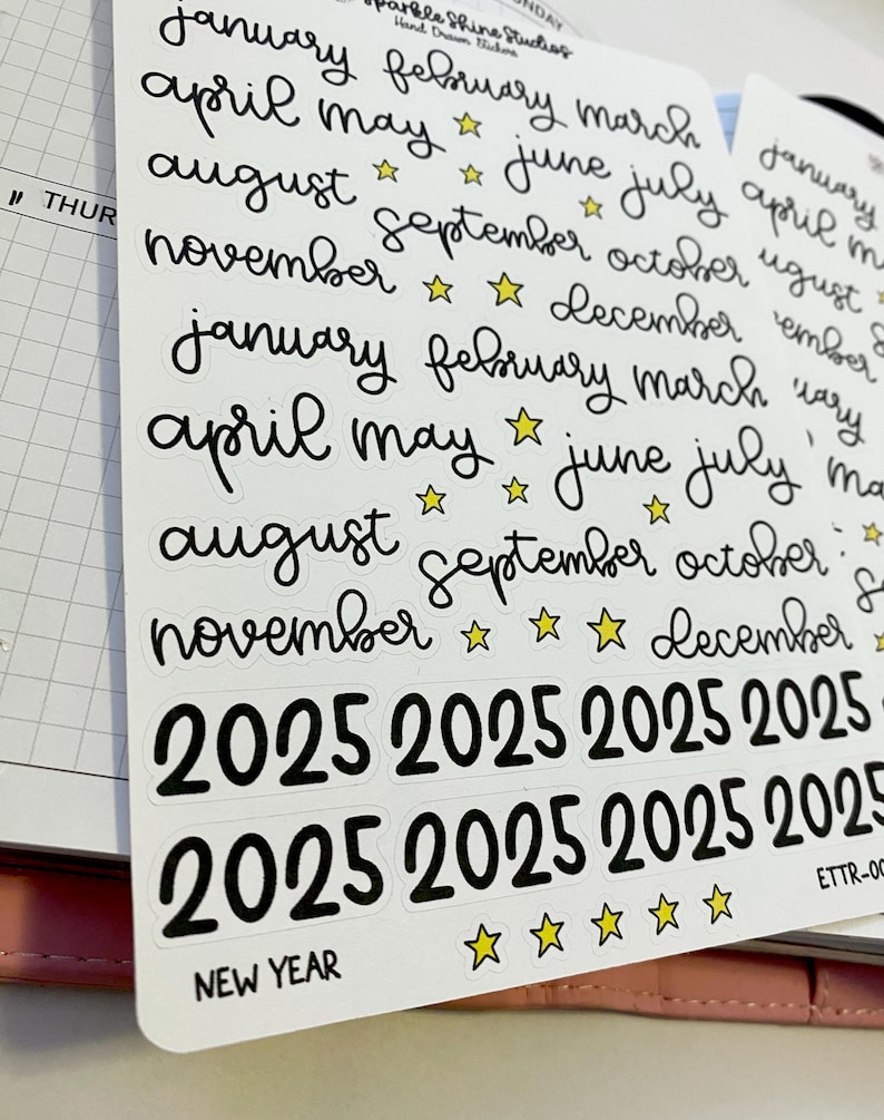 May include: A sheet of white sticker paper with black handwritten text and yellow star stickers. The stickers include the months of the year and the year 2025. The text "NEW YEAR" is also included on the sheet.