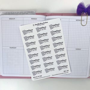 May include: A sheet of white stickers with the text "replace AC filters" in black script and a small gray air filter graphic. The stickers are on a pink planner, with a purple bow and gold paper clip.