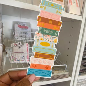 以下が含まれることがあります： 花柄と幾何学模様のカラフルな本のステッカーのセット。ステッカーは縦に積み重ねられており、一番上のステッカーは金色がかった薄い青色の本です。これらのステッカーは、プランナー、ジャーナル、スクラップブックに遊び心のあるアクセントを加えるのに最適です。