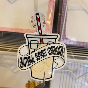 Può includere: Un adesivo bianco con un'illustrazione di cartone animato di un bicchiere di limonata con una cannuccia a strisce rosse e bianche. Il testo "Emotional Support Lemonade" è scritto su uno striscione nero attorno al bicchiere.