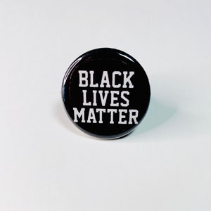 Peut inclure: Épinglette bouton noire avec le texte "Black Lives Matter" en lettres blanches sur fond noir.