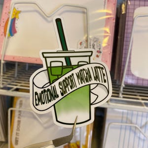 Può includere: Un adesivo verde e bianco con un'immagine di cartone animato di una tazza di latte matcha con una cannuccia e il testo "Emotional Support Matcha Latte".