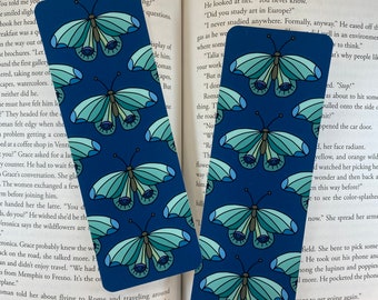 Puede incluir: Un marcapáginas azul con un patrón repetitivo de mariposas con alas azules, verdes y blancas y ojos grandes. Las mariposas están sobre un fondo azul oscuro.