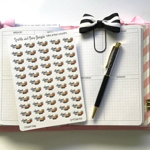 May include: A planner spread with a sheet of "Cheat Day" stickers featuring burgers and fries. A black and white striped bow clip and a pen with a glittery barrel are also visible. The planner is open to a grid-lined page.