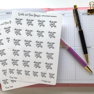 May include: Two sheets of white sticker paper with the phrase "This Too Shall Pass" repeated in black cursive writing. The stickers are arranged in a grid pattern. The sheets are on a pink and white planner.