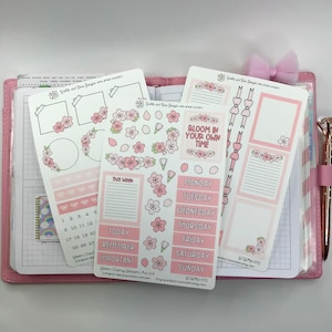 以下が含まれることがあります： 桜のテーマのピンクと白のプランナー用ステッカーのセット。ステッカーには、カレンダー、デイリープランナー、ウィークリープランナー、さまざまな装飾要素が含まれています。ステッカーは、プランナーに遊び心を加えるのに最適です。