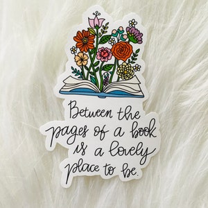 Può includere: Un adesivo raffigurante un libro aperto con fiori colorati che sbocciano dalle sue pagine. L'adesivo presenta il testo "Between the pages of a book is a lovely place to be" in un carattere scritto a mano.