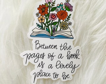 Può includere: Un adesivo raffigurante un libro aperto con fiori colorati che sbocciano dalle sue pagine. L'adesivo presenta il testo "Between the pages of a book is a lovely place to be" in un carattere scritto a mano.