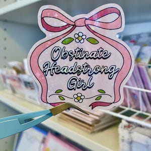 Può includere: Un adesivo rosa con le parole "Obstinate Headstrong Girl" in un carattere decorativo. L'adesivo ha un fiocco in alto e accenti floreali. L'adesivo è tenuto da un paio di pinzette azzurre.