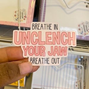Può includere: Un adesivo bianco con la scritta "Breathe In Unclench Your Jaw Breathe Out" in lettere rosa. L'adesivo è tenuto in mano, con altri oggetti sullo sfondo.
