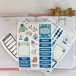 May include: A set of winter-themed planner stickers with blue, white, and gray accents. The stickers include a snow globe, mittens, a hat, a snowman, and a calendar with the days of the week. The stickers are on a pink and white planner.