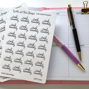 May include: A sheet of white sticker paper with the text "Work Remotely" repeated multiple times in black cursive writing. The sticker sheet is on top of a pink and white gridded planner.