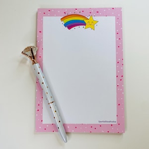 May include: Pink notepad with a white interior and a rainbow shooting star with a smiling face. The notepad has a pink border with white polka dots. The text "SparkleShineStudio" is printed in the bottom right corner.
