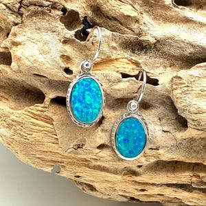 May include: A pair of silver-toned earrings with oval-shaped, vibrant blue opal stones. The earrings have a textured silver border and a hook closure. The opals display a mix of blue and turquoise hues, creating a striking contrast against the silver.