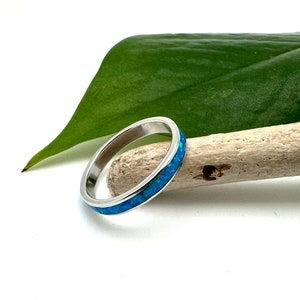 Puede incluir: Un anillo de plata con una banda de ópalo azul incrustado. El anillo está sobre un fondo blanco, con una hoja verde y un trozo de madera flotante en el fondo. El anillo tiene un diseño sencillo y elegante.