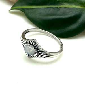 May include: A silver ring featuring a sunburst design encircling a pale blue, oval gemstone. The ring is set against a white backdrop, with a green leaf and a piece of wood in the background. This is a piece of jewellery.