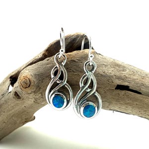 May include: A pair of silver-toned drop earrings with a swirling design. Each earring features a round, blue gemstone at the bottom. The earrings are displayed on a piece of driftwood against a white background.