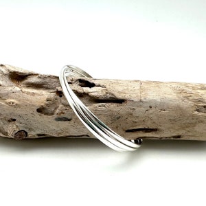 May include: A silver bangle bracelet with three bands, resting on a piece of driftwood.