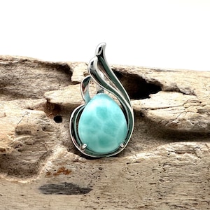 May include: A teardrop-shaped pendant with a light blue gemstone, set in a silver-colored metal frame. The frame has a flowing, organic design. The pendant is displayed on a piece of driftwood.