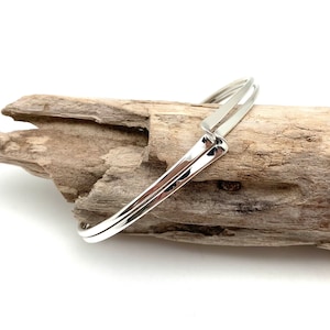May include: A silver bangle bracelet with a simple, modern design. The bracelet is made of two thin bands of metal that are joined together at the ends. The bracelet is shown on a piece of driftwood.