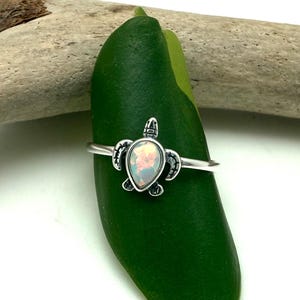 Puede incluir: Anillo de plata con diseño de tortuga. La tortuga tiene una piedra tipo ópalo en su caparazón. El anillo se muestra sobre una hoja verde, con un trozo de madera flotante en el fondo. Joyería.
