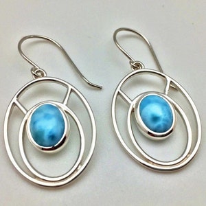 K&ouml;nnte beinhalten: Ein Paar silberne Ohrringe mit einem blauen ovalen Edelstein, der in einem runden Rahmen gefasst ist. Die Ohrringe haben ein schlichtes, modernes Design.