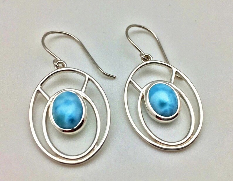 K&ouml;nnte beinhalten: Ein Paar silberne Ohrringe mit einem blauen ovalen Edelstein, der in einem runden Rahmen gefasst ist. Die Ohrringe haben ein schlichtes, modernes Design.