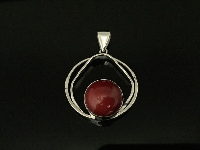 Pu&ograve; includere: Un ciondolo d'argento con una pietra preziosa rossa incastonata in un design a doppio cerchio.