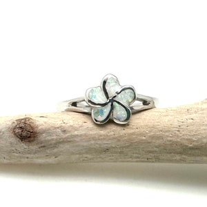 Può includere: Un anello d'argento con un designo a forma di fiore di opale bianco. L'anello è su un pezzo di legno alla deriva.