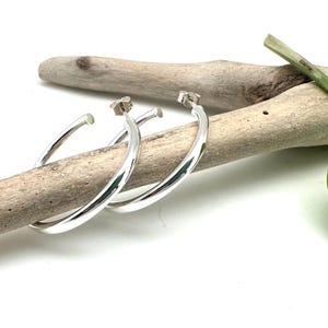 May include: A pair of silver hoop earrings with a smooth, rounded finish. The earrings are displayed on a piece of driftwood with a green leaf.