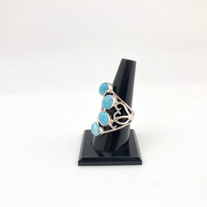 May include: A silver ring with a delicate filigree design and five light blue gemstones. The ring is displayed on a black ring stand.