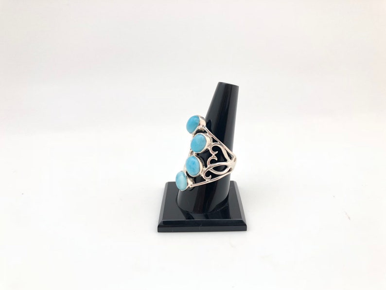 May include: A silver ring with a delicate filigree design and five light blue gemstones. The ring is displayed on a black ring stand.