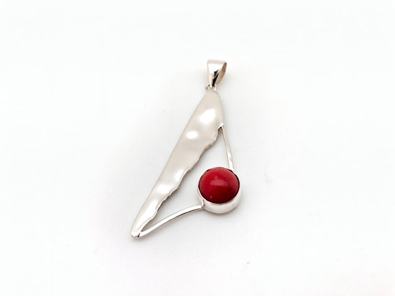 May include: A silver pendant with a red coral stone in the center. The pendant is shaped like a triangle with a curved edge.