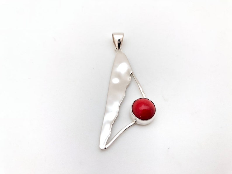 May include: A silver pendant with a red coral stone in the center. The pendant is shaped like a triangle with a curved, textured edge.
