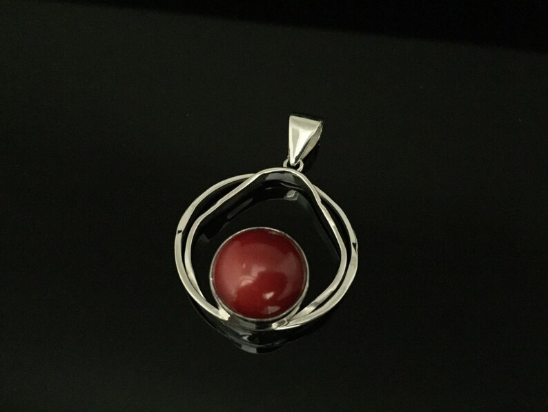 Pu&ograve; includere: Un ciondolo d'argento con una pietra preziosa rossa incastonata in un design a doppio cerchio.