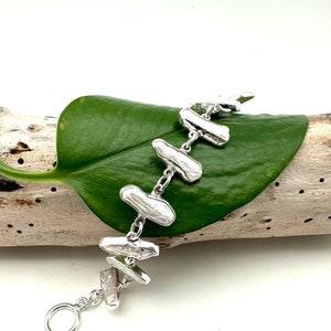 Puede incluir: Un brazalete de plata con una cadena y varios abalorios de perlas blancas de forma irregular. El brazalete se muestra sobre un trozo de madera a la deriva con una hoja verde.