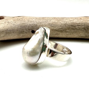May include: A silver ring with a large, white, pearlescent gemstone. The ring is set on a piece of driftwood.
