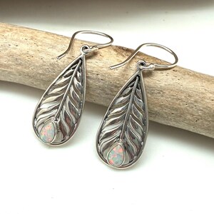 May include: A pair of silver-toned drop earrings with a feather design. Each earring features a teardrop shape with detailed feather etching and a small, oval-shaped opal stone at the bottom. The earrings are displayed on a light-colored wooden surface.