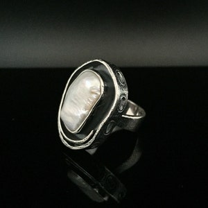 May include: A silver ring with a large, white, oval-shaped pearl set in a black, textured metal bezel.