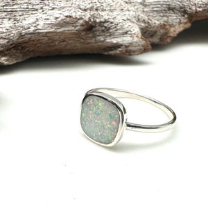 May include: A silver ring with a square, iridescent opal stone. The opal displays a mix of green, blue, and orange hues. The ring is set on a white surface, with a piece of driftwood in the background.