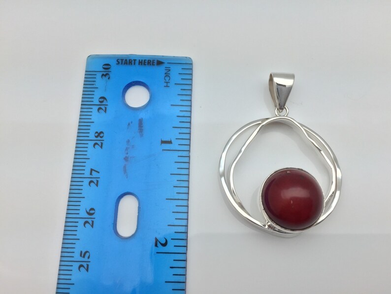 Pu&ograve; includere: Ciondolo in argento con una pietra preziosa rossa incastonata in un design a doppio cerchio. Il ciondolo misura circa 2,5 cm di diametro.
