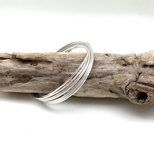 May include: A silver bangle bracelet made of multiple thin bands of metal. The bracelet is lying on a piece of driftwood.