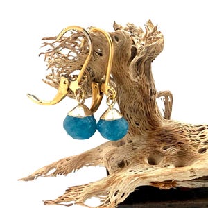 May include: A pair of gold-colored leverback earrings with faceted, light blue gemstone drops. The gemstones are capped with gold-colored metal. The earrings are displayed on a piece of textured, light brown wood.