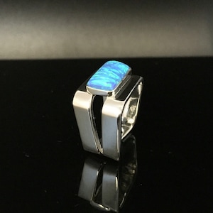 Puede incluir: Un anillo de plata con una piedra preciosa de ópalo azul engastada en una banda de forma cuadrada.