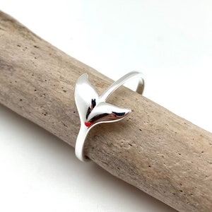 Könnte beinhalten: Ein silberner Ring mit einem Wal-Schwanz-Design. Der Ring liegt auf einem Stück Treibholz.